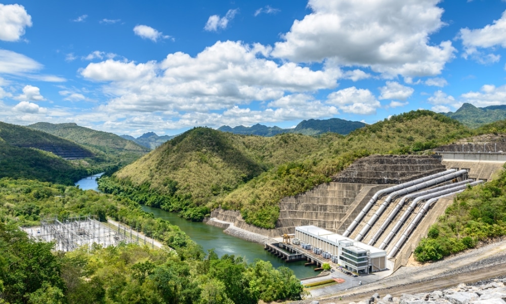 โรงไฟฟ้าพลังน้ำที่มีท่อขนาดใหญ่ตั้งอยู่บนเนินเขาข้างแม่น้ำ ล้อมรอบด้วยเนินเขาสีเขียวและท้องฟ้าที่มีเมฆบางส่วน—เป็นหนึ่งในสถานที่ท่องเที่ยวที่โดดเด่นของกาญจนบุรีสำหรับผู้ที่รักธรรมชาติและสิ่งมหัศจรรย์ทางวิศวกรรม.