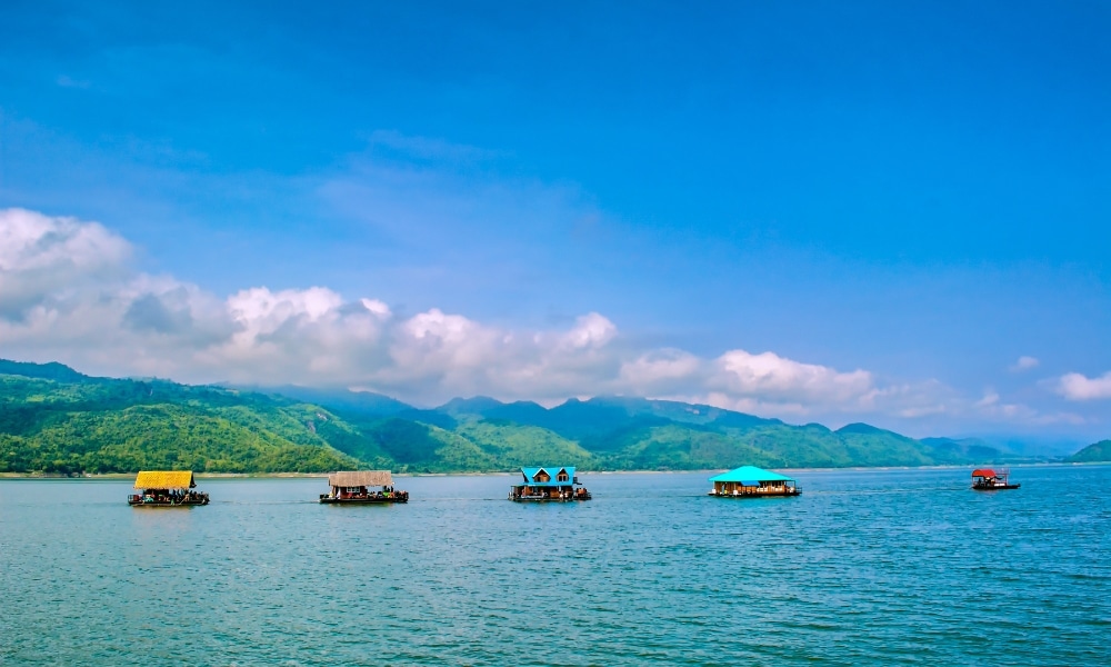 บ้านลอยน้ำที่มีหลังคาสีสันสดใสกระจายอยู่ทั่วทะเลสาบที่เงียบสงบ ล้อมรอบด้วยเนินเขาสีเขียวและท้องฟ้าสีฟ้าที่มีเมฆบางส่วน—เป็นภาพอันเป็นเอกลักษณ์ของสถานที่ท่องเที่ยวในกาญจนบุรีที่เน้นเสน่ห์ของสถานที่ท่องเที่ยวในกาญจนบุรี.