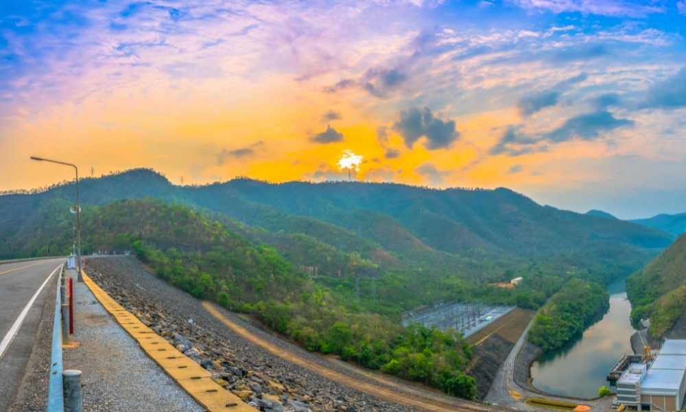 ถนนลาดยางทอดยาวขนานไปกับเขื่อนขนาดใหญ่ที่มีน้ำล้อมรอบ ภูเขาเขียวขจีและป่าไม้ทอดตัวอยู่ใต้ท้องฟ้าสีสันสดใสยามพระอาทิตย์ตกดิน—สถานที่ที่เหมาะสำหรับการท่องเที่ยวในกาญจนบุรี.