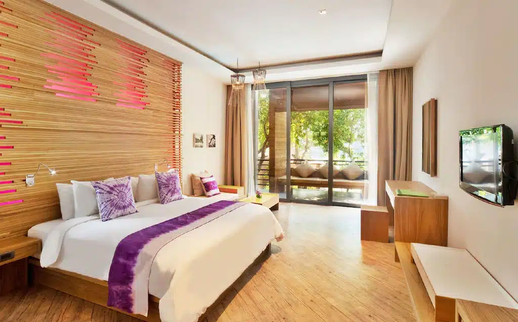 ห้องพักโรงแรมสไตล์ทันสมัย พร้อมเตียงคิงไซส์ รายละเอียดตกแต่งสีม่วง ผนังตกแต่งด้วยไม้แผ่น ทีวีจอแบน และหน้าต่างขนาดใหญ่ที่มองเห็นวิวต้นไม้และระเบียง — เหมาะอย่างยิ่งสำหรับผู้ที่มองหาที่พักเกาะเสม็ดติดทะเล ที่ผสมผสานความสะดวกสบายและสไตล์ได้อย่างลงตัว.
