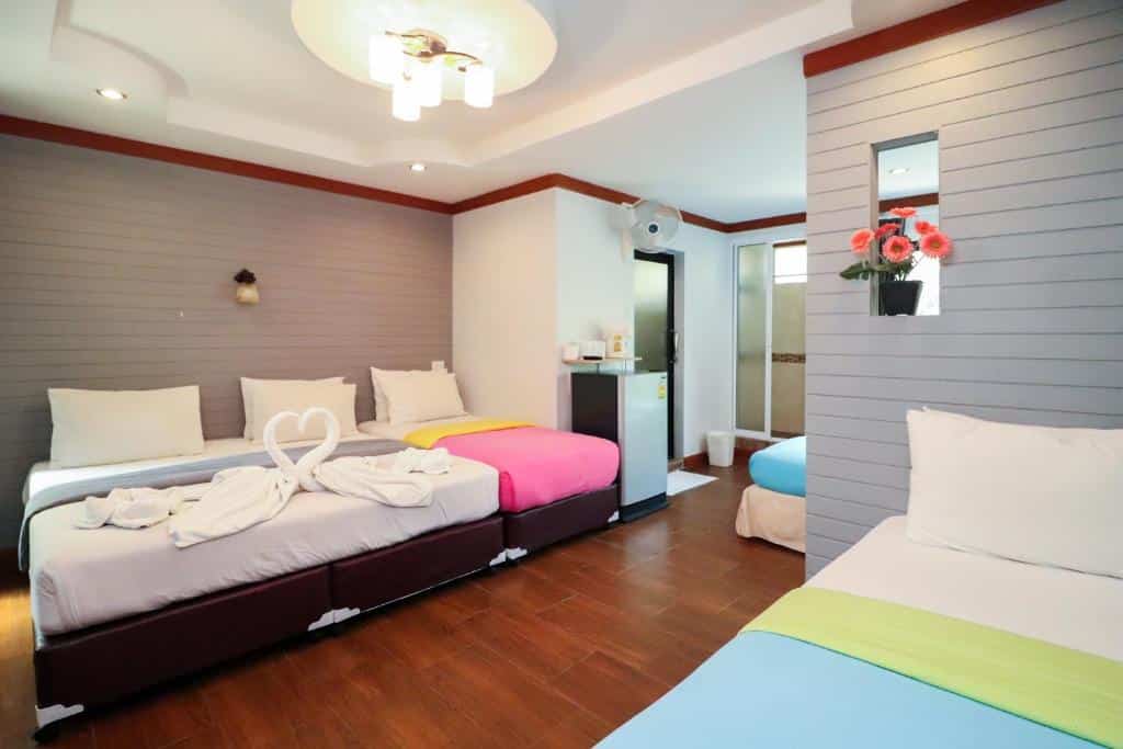 ห้องพักในโรงแรมที่มีเตียงสามเตียง ชุดเครื่องนอนสีสันสดใส ผ้าขนหนูพับเป็นรูปหงส์บนเตียงหนึ่ง ดอกไม้ประดับบนผนัง พื้นไม้ และระบบไฟส่องสว่างแบบทันสมัย — เป็นตัวเลือกที่สมบูรณ์แบบสำหรับผู้ที่กำลังมองหา ที่พักเกาะเสม็ดติดทะเล เพื่อการพักผ่อนที่สะดวกสบายและน่าจดจำ.