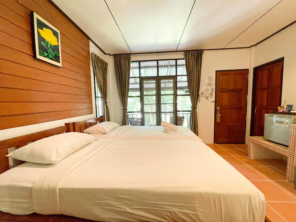ห้องพักในโรงแรมที่มีเตียงสองเตียงจัดเรียงอย่างเรียบร้อย ตู้เย็นขนาดเล็ก ผนังไม้ที่มีภาพวาดดอกไม้สีเหลือง ม่าน และประตูที่นำไปสู่ระเบียง ซึ่งสามารถมองเห็นต้นไม้ด้านนอกได้ — เหมาะอย่างยิ่งสำหรับผู้ที่กำลังมองหาที่พักเกาะเสม็ดติดทะเล.