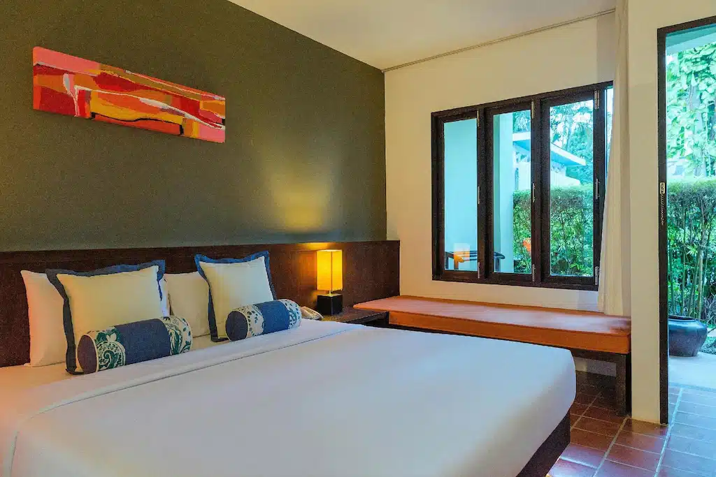 ห้องพักในโรงแรมที่มีเตียงขนาดใหญ่ หมอนตกแต่ง โคมไฟข้างเตียง ภาพวาดบนผนัง และที่นั่งริมหน้าต่างที่อบอุ่น ซึ่งสามารถมองเห็นความเขียวชอุ่มของธรรมชาติด้านนอกได้อย่างชัดเจน — เหมาะอย่างยิ่งสำหรับแขกที่กำลังมองหาที่พักบนเกาะเสม็ด ระหว่างการพักผ่อนบนเกาะ.