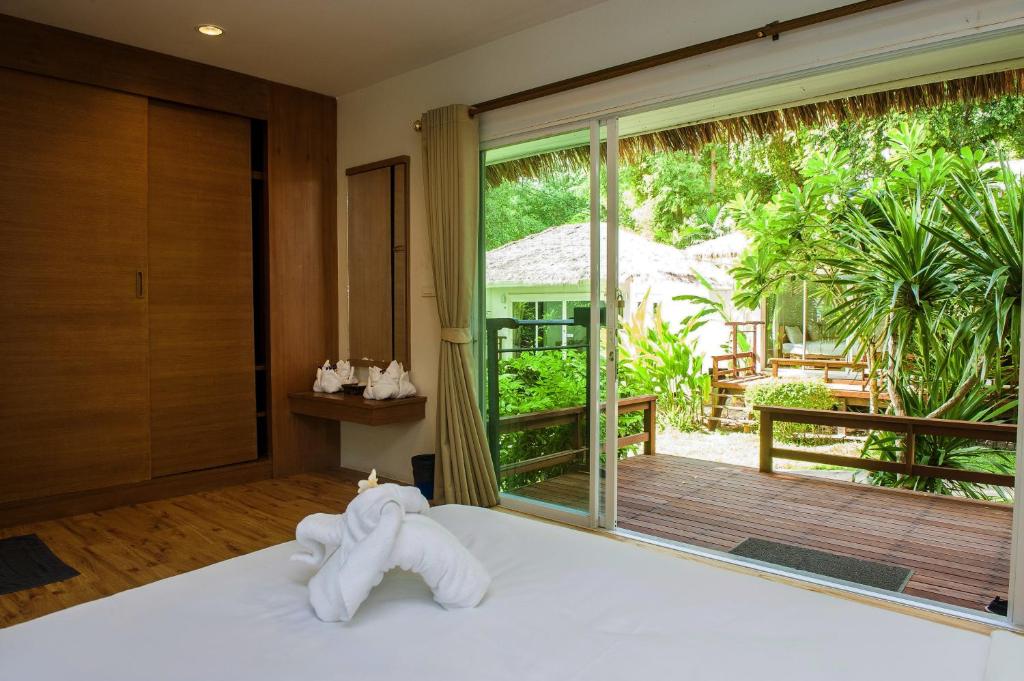 ห้องพักในโรงแรมที่มีพื้นไม้และประตูเลื่อนกระจกที่เปิดออกสู่ระเบียงซึ่งมีพืชพรรณเขียวชอุ่มและพืชเขตร้อนอยู่ด้านนอก เหมาะอย่างยิ่งสำหรับผู้ที่กำลังมองหาที่พักเกาะเสม็ดติดทะเล พื้นที่อันน่าดึงดูดนี้มีเตียงที่ตกแต่งด้วยผ้าขนหนูพับเป็นรูปช้าง มอบประสบการณ์พักผ่อนอย่างสงบให้กับแขกผู้เข้าพัก เพียงไม่กี่ก้าวจากความงามของชายหาดเกาะเสม็ด.