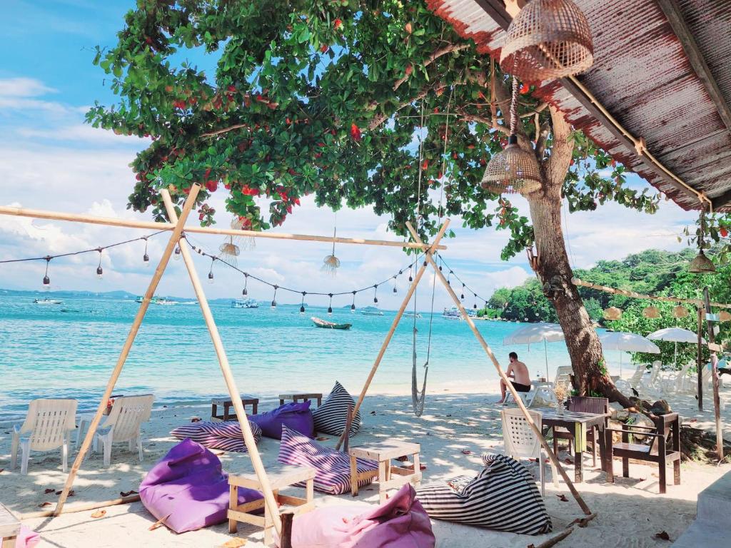พื้นที่นั่งเล่นริมชายหาดที่มีเบาะนั่งแบบบีนแบ็ก ชิงช้า โต๊ะ และเก้าอี้ใต้ต้นไม้ข้างทะเลในวันที่มีแดดสดใส; เป็นสถานที่ที่สมบูรณ์แบบสำหรับการพักผ่อนที่ที่พักเกาะเสม็ดติดทะเล โดยมีเพียงไม่กี่คนที่กำลังผ่อนคลาย ขณะที่เรือลำเล็กแล่นผ่านไปมาในระยะไกล.