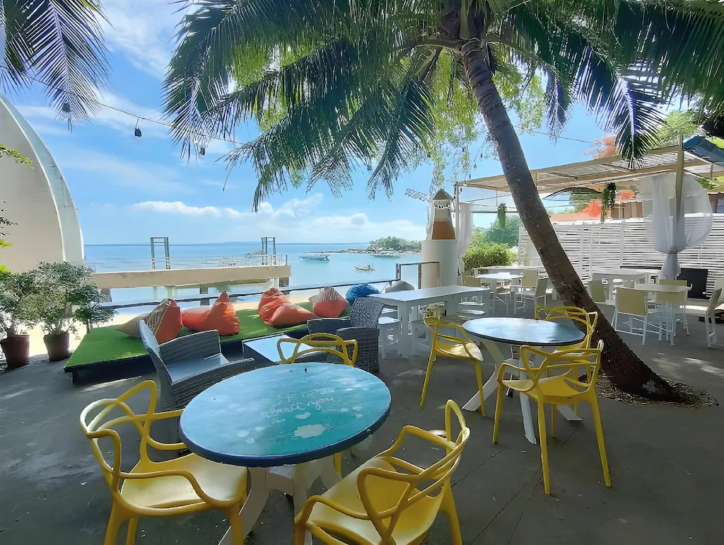 คาเฟ่กลางแจ้งที่มีเก้าอี้สีเหลืองและโต๊ะกลมใต้ต้นปาล์ม มองออกไปเห็นชายหาดและทะเล พร้อมด้วยเบาะนั่งแบบบีนแบ็ก และท้องฟ้าสีฟ้าใสเป็นฉากหลัง — เหมาะอย่างยิ่งสำหรับการพักผ่อนที่ที่พักเกาะเสม็ดติดทะเล ซึ่งคุณสามารถเพลิดเพลินกับวิวชายทะเลที่สวยงามได้เพียงไม่กี่ก้าวจากห้องพักของคุณ.
