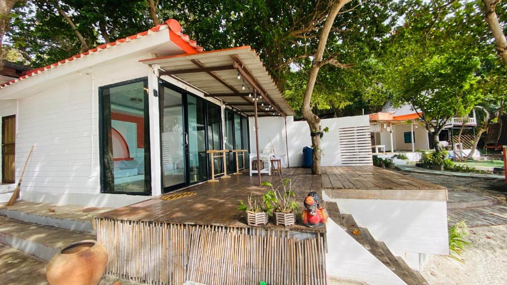 บ้านบังกะโลสีขาวสไตล์โมเดิร์น พร้อมประตูแก้วขนาดใหญ่ ระเบียงไม้ ต้นไม้ในกระถาง และต้นไม้ที่ให้ร่มเงา ตั้งอยู่บนพื้นที่ทรายใกล้กับอาคารอื่นๆ ที่มีลักษณะคล้ายกัน — เหมาะอย่างยิ่งสำหรับผู้ที่กำลังมองหา ที่พักเกาะเสม็ดติดทะเล เพื่อพักผ่อนอย่างสบายใจ.