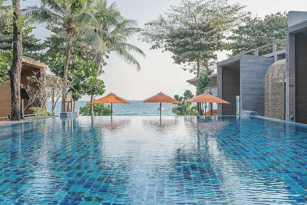 สระว่ายน้ำแบบอินฟินิตี้ที่ปูกระเบื้องสีฟ้า มีร่มสีส้มสามคันตั้งอยู่ในน้ำ พร้อมด้วยต้นปาล์ม และวิวทะเลเป็นฉากหลัง ล้อมรอบด้วยอาคารสมัยใหม่ — ที่พักเกาะเสม็ดติดทะเลที่สมบูรณ์แบบสำหรับการพักผ่อนอย่างสบายใจ.