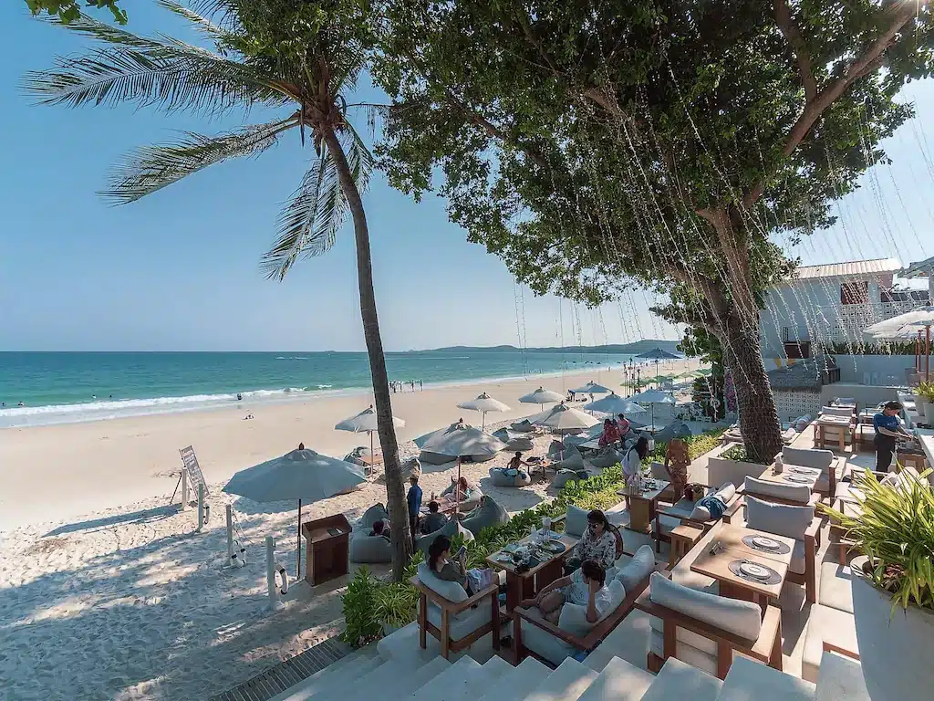 พื้นที่นั่งรับประทานอาหารกลางแจ้งของร้านอาหารมีวิวที่มองเห็นชายหาดทรายขาวพร้อมร่มชายหาดและผู้คนกำลังพักผ่อน ต้นปาล์มและไฟประดับช่วยสร้างร่มเงาและตกแต่งบรรยากาศใต้ท้องฟ้าสีฟ้าใส สร้างบรรยากาศที่สมบูรณ์แบบสำหรับแขกที่กำลังมองหา ที่พักเกาะเสม็ดติดทะเล หรือ ที่พักเสม็ด เพื่อผ่อนคลายและเพลิดเพลินกับวิวชายฝั่งที่สงบเงียบ.