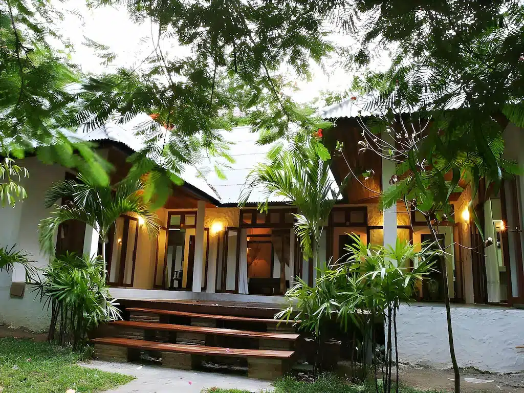 บ้านสีขาวที่มีขอบไม้และหลังคาสังกะสี ล้อมรอบด้วยพืชพรรณเขียวชอุ่มและต้นปาล์มเล็กๆ ให้ความรู้สึกอบอุ่นต้อนรับด้วยบันไดที่นำไปสู่ระเบียงและแสงไฟที่ส่องสว่างจากภายใน — เป็นตัวเลือกที่สมบูรณ์แบบสำหรับผู้ที่กำลังมองหา ที่พักเกาะเสม็ด หรือใครก็ตามที่กำลังมองหา เกาะเสม็ดที่พัก ที่มีเอกลักษณ์และตั้งอยู่ท่ามกลางธรรมชาติ.