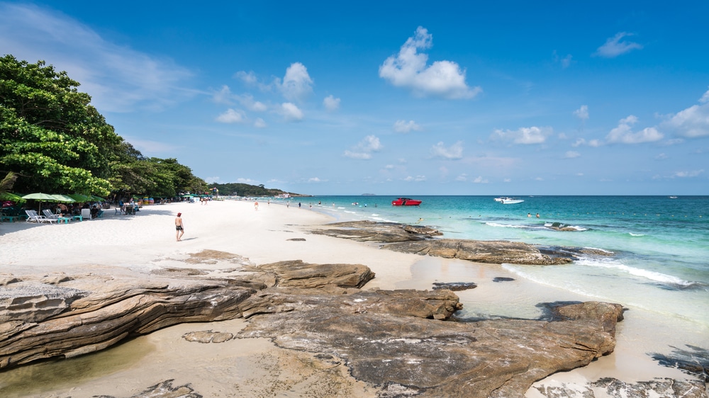 ชายหาดทรายขาวกับชายฝั่งหิน ผู้คนกำลังเดินเล่น ต้นไม้เขียวขจีทางซ้าย น้ำทะเลสีเทอร์ควอยซ์ เรือหลายลำในระยะไกล และท้องฟ้าสีฟ้าที่มีเมฆกระจายตัว—ภาพที่งดงามราวกับสวรรค์ เหมาะอย่างยิ่งสำหรับผู้ที่กำลังมองหา ที่พักเกาะเสม็ดติดทะเล หรือที่พักบนเกาะเสม็ดที่ผ่อนคลาย ซึ่งอยู่เพียงไม่กี่ก้าวจากชายหาด.