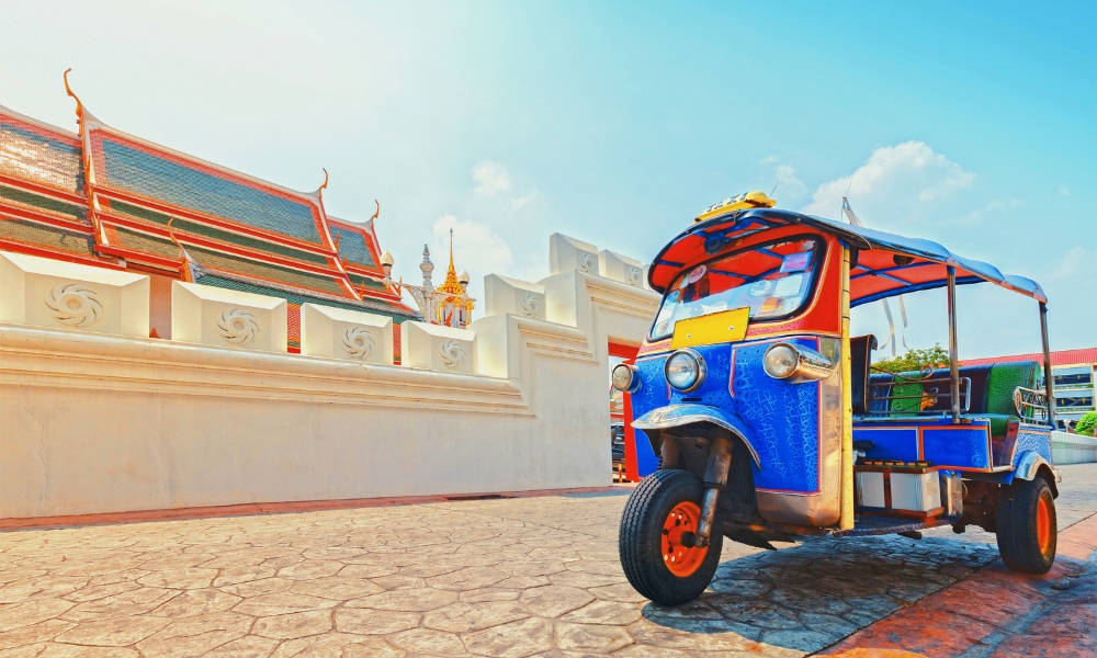 รถตุ๊กตุ๊กสีฟ้าจอดอยู่บนถนนลาดยางข้างวัดไทยแบบดั้งเดิมที่มีรายละเอียดหลังคาอันวิจิตรภายใต้ท้องฟ้าสีฟ้าใส ถ่ายทอดเสน่ห์ของสถานที่ท่องเที่ยวในกรุงเทพฯ.