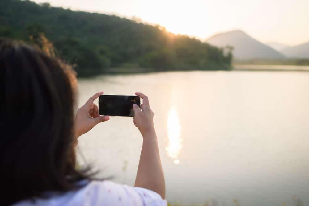 ผู้หญิงกำลังถ่ายภาพทิวทัศน์อันงดงามด้วยโทรศัพท์มือถือของเธอที่ทะเลสาบอันงดงามใน เที่ยวสุโขทัย
