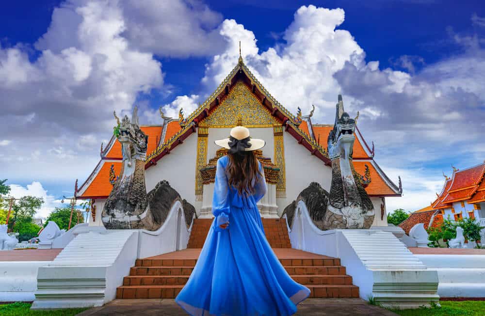 ผู้หญิงสวมชุดสีฟ้าและหมวก ยืนหันหน้าสู่วัดไทยแบบดั้งเดิมที่มีรายละเอียดประดับตกแต่งอย่างวิจิตร พร้อมด้วยเมฆที่ดูน่าทึ่งบนท้องฟ้า และรูปปั้นมังกรสองตัวตั้งอยู่ทั้งสองข้างทางเข้า — เป็นภาพที่โดดเด่นสำหรับผู้ที่กำลังมองหา ที่เที่ยวน่าน เพื่อสัมผัสเสน่ห์ทางวัฒนธรรมของภูมิภาคที่สวยงามแห่งนี้อย่างเต็มที่.