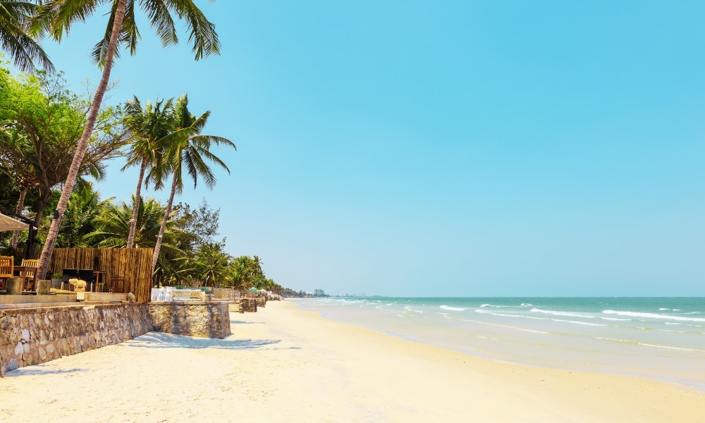 ชายหาดทรายขาวละเอียดพร้อมคลื่นน้ำทะเลอ่อนโยน ท้องฟ้าสีครามใส และต้นปาล์มเรียงรายอยู่ริมกำแพงหินและรั้วไม้ทางด้านซ้าย—เหมาะอย่างยิ่งสำหรับผู้ที่ต้องการสัมผัสประสบการณ์ที่พักในปราณบุรีที่เงียบสงบ.