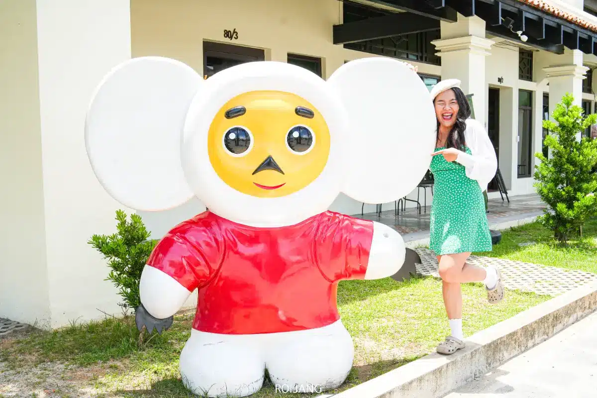 ผู้หญิงในชุดสีเขียวโพสท่าเล่น ๆ ข้าง ๆ รูปปั้นตัวการ์ตูนขนาดใหญ่ที่มีหูใหญ่ เสื้อสีแดง และใบหน้าสีเหลืองนอกอาคาร Spice House.