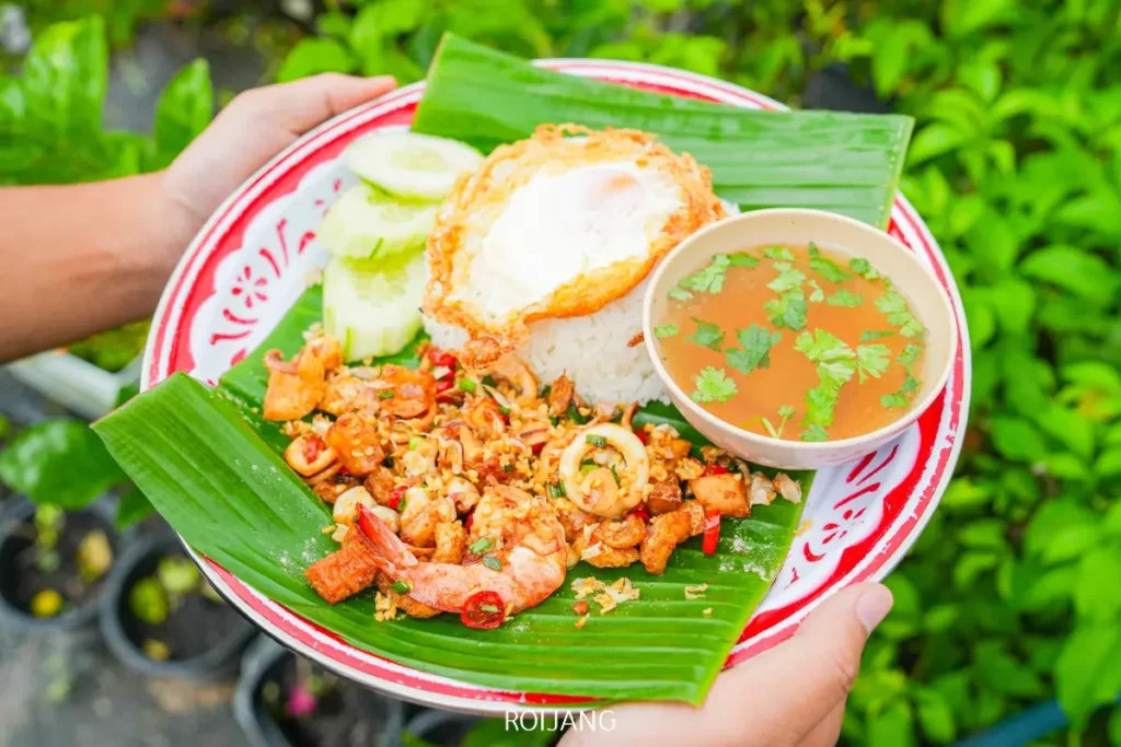 มือหนึ่งถือจานข้าวที่โรยด้วยไข่ดาว แตงกวาหั่นบาง อาหารทะเลผัดจากร้าน ลุงป้า กะเพราถาด และชามซุปใส ทั้งหมดจัดวางอย่างสวยงามบนใบตอง.