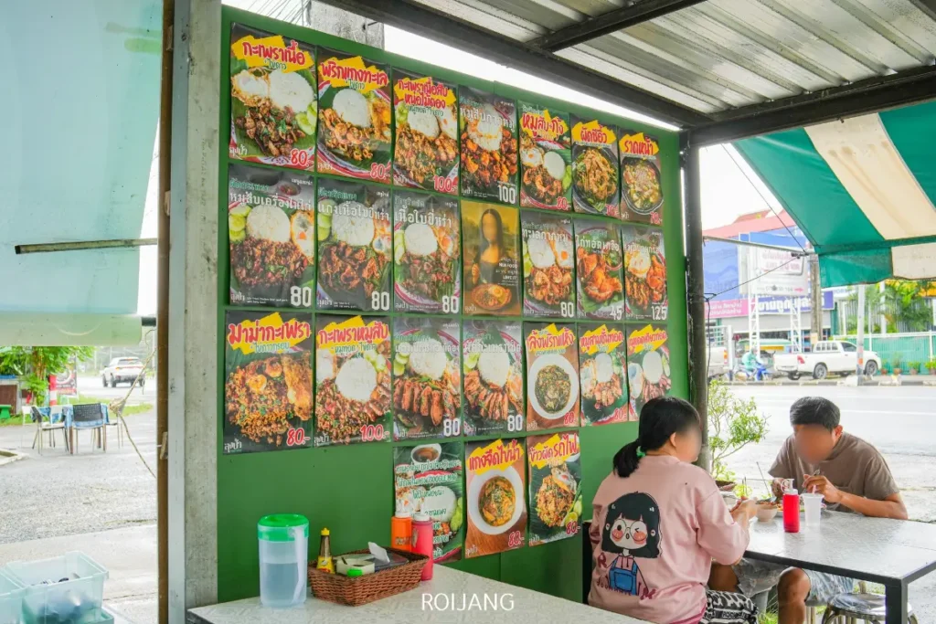 สองคนนั่งอยู่ที่โต๊ะในร้านอาหารกลางแจ้ง โดยมีผนังอยู่เบื้องหลังพวกเขาซึ่งแสดงรูปภาพเมนูอาหารไทยสีสันสดใสและราคาจาก ลุงป้า กะเพราถาด.