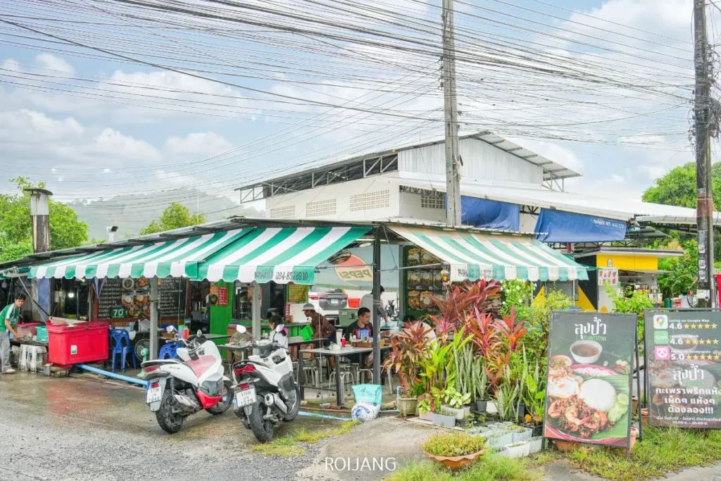 ร้านอาหารริมถนน ลุงป้า กะเพราถาด มีร่มกันแดดลายทางสีเขียวและขาว จอดรถจักรยานยนต์ไว้ ติดป้ายเมนู มีต้นไม้เขียวชอุ่ม และผู้คนหลายคนที่กำลังรับประทานอาหารกลางแจ้งใต้ท้องฟ้าที่มีเมฆครึ้ม.