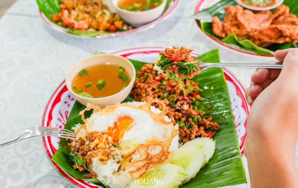 จานจาก ลุงป้า กะเพราถาด ประกอบด้วยข้าว, ไข่ดาว, เนื้อสับผัด, แตงกวาหั่น, และซุปบนใบตอง มือยกส้อมที่มีอาหารอยู่เหนือจาน.