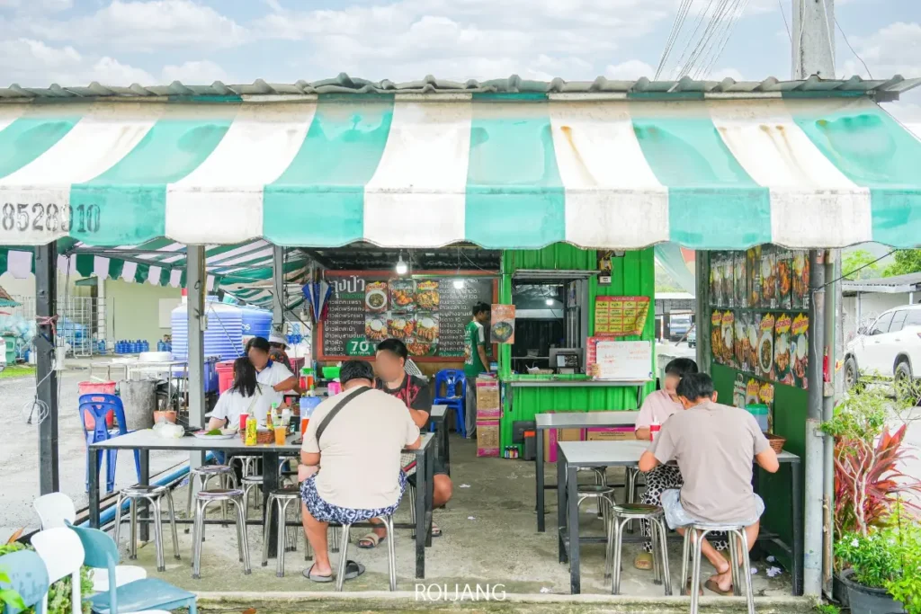 ร้านขายอาหารริมทางกลางแจ้ง ลุงป้ากะเพราถาด มีร่มกันแดดลายทางสีเขียวและขาว คนหลายนั่งอยู่ที่โต๊ะกำลังเพลิดเพลินกับอาหาร และมีป้ายเมนูแสดงไว้ที่ร้าน.