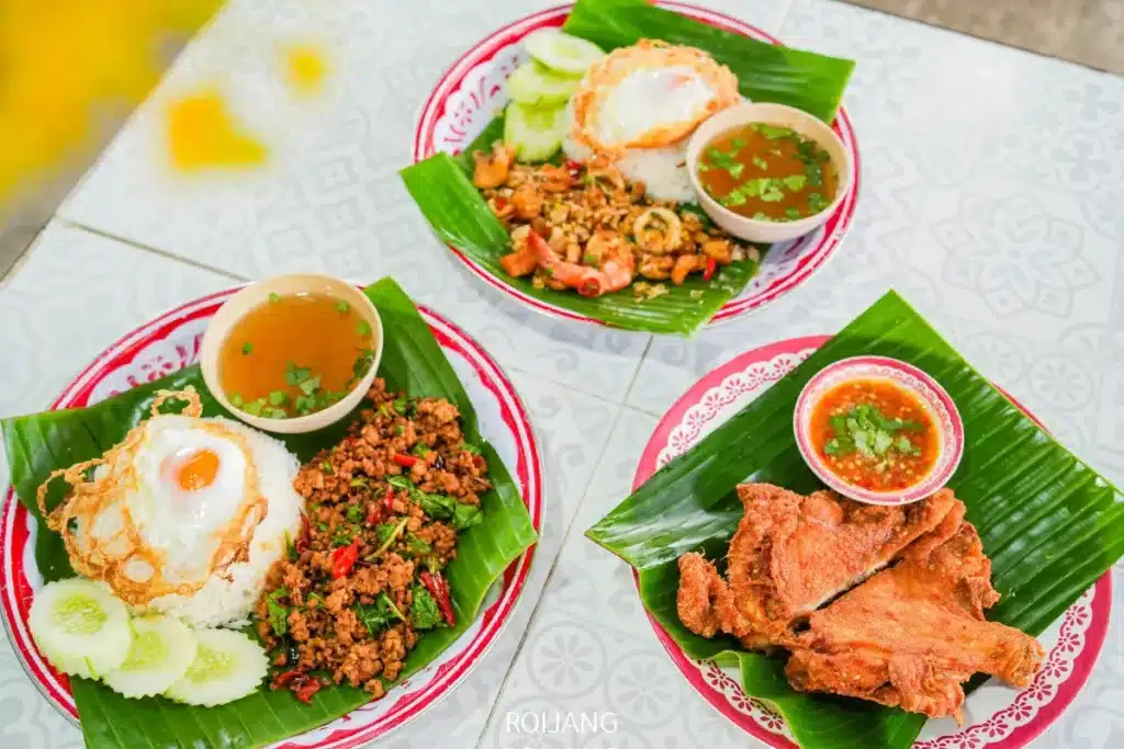 อาหารไทยสามจานจากร้าน ลุงป้า กะเพราถาด บนโต๊ะ: สองจานมีข้าว ไข่ดาว อาหารผัด และซุป และอีกหนึ่งจานมีไก่ทอดกับน้ำจิ้ม ทั้งหมดเสิร์ฟบนใบตอง.