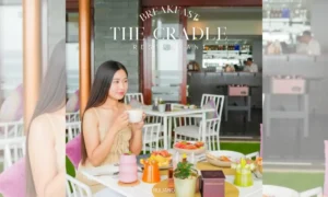 ผู้หญิงคนหนึ่งนั่งอยู่ที่โต๊ะในร้านอาหารชื่อ The Cradle ล้อมรอบด้วยอาหารเช้าหลากหลายชนิดและเครื่องดื่ม เธอกำลังถือถ้วยในมือและมองไปทางข้างๆ บรรยากาศสว่างไสวและทันสมัย.
