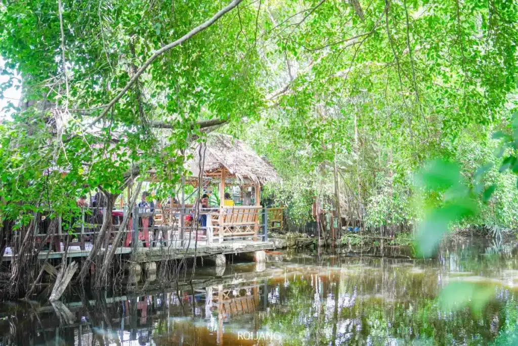 กระท่อมไม้หลังคามุงจากตั้งอยู่บนเสาสูงเหนือผืนน้ำสงบ รายล้อมไปด้วยพุ่มไม้เขียวชอุ่มและต้นไม้ใหญ่ ชวนให้คุณมาลิ้มลอง ทองปาล์มก๋วยเตี๋ยวต้มยำน้ำข้น ในบรรยากาศธรรมชาติอันเงียบสงบ.