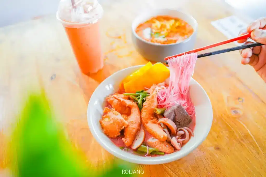 ชามหนึ่งมีก๋วยเตี๋ยวต้มยำน้ำข้นใส่หน่อไม้ฝรั่งทะเลและผักวางอยู่บนโต๊ะไม้ ขณะที่อีกชามหนึ่งเป็นซุป และแก้วพลาสติกใส่น้ำส้มวางอยู่ข้างกัน มือหนึ่งกำลังยกเส้นก๋วยเตี๋ยวด้วยตะเกียบ.