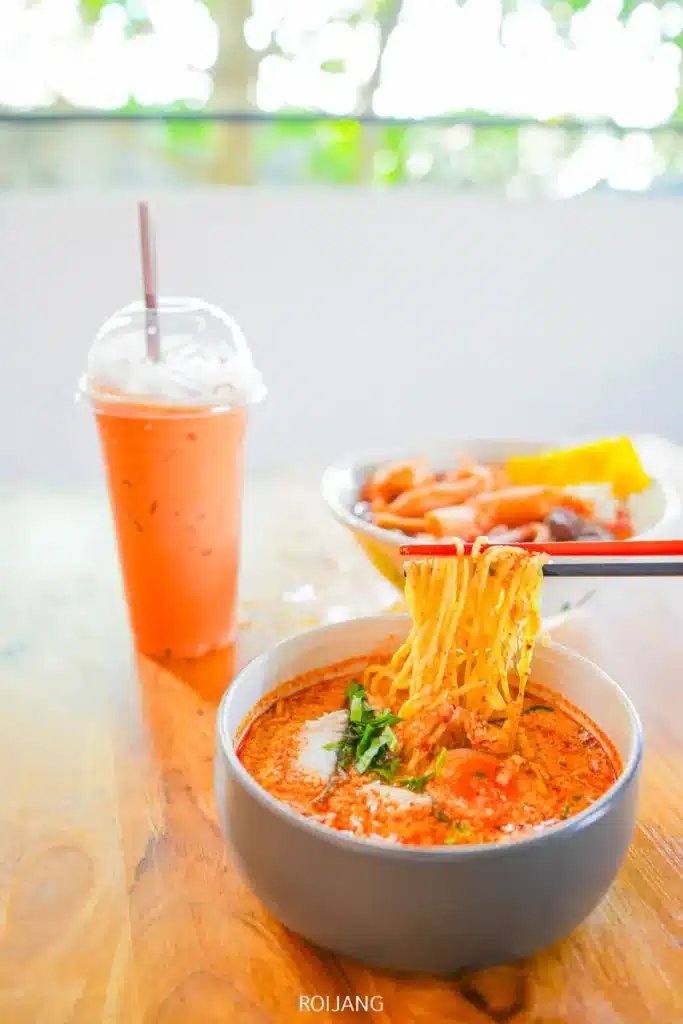 ชามก๋วยเตี๋ยวต้มยำน้ำข้น ทองปาล์ม รสเผ็ด พร้อมตะเกียบคีบเส้น ชามเครื่องเคียงหลากหลาย และเครื่องดื่มเย็นบนโต๊ะไม้.