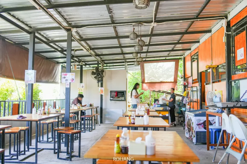 ร้านอาหารกลางแจ้งแบบสบายๆ มีโต๊ะและม้านั่งไม้ ขวดเครื่องปรุงบนโต๊ะ และผู้คนไม่กี่คนที่กำลังรับประทานอาหารและทำงานในพื้นที่ที่สว่างและมีหลังคาคลุม เสิร์ฟ ทองปาล์มก๋วยเตี๋ยวต้มยำน้ำข้น.