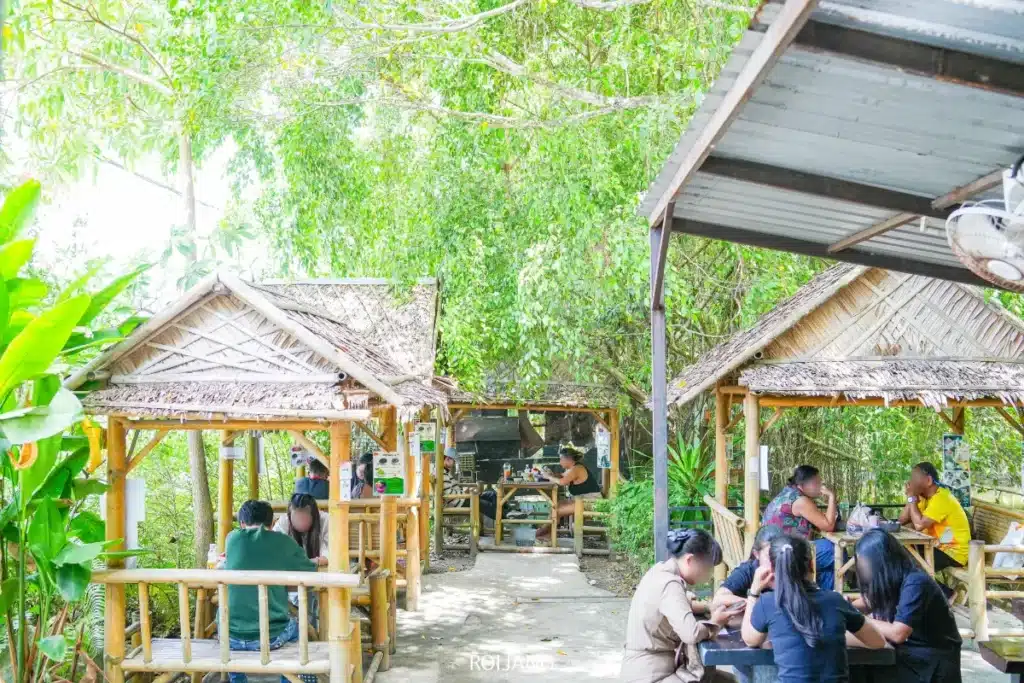 ผู้คนนั่งรับประทานอาหารที่โต๊ะไม้ใต้กระท่อมหลังคามุงจาก เพลิดเพลินกับก๋วยเตี๋ยวต้มยำน้ำข้นรสจัด เสิร์ฟพร้อมใบตอง ท่ามกลางบรรยากาศเขียวขจีในวันที่แดดสดใส.