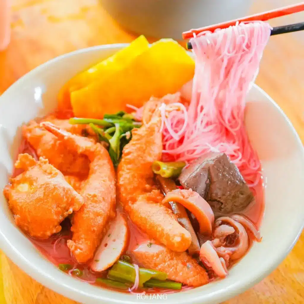 ชามก๋วยเตี๋ยวต้มยำน้ำข้นสีชมพู ทองปาล์ม ก๋วยเตี๋ยวต้มยำน้ำข้น พร้อมลูกชิ้นปลา น่องไก่ ผักสด ผักดองสีเหลือง และเลือดหมูต้มแข็ง พร้อมตะเกียบคีบเส้นก๋วยเตี๋ยว.