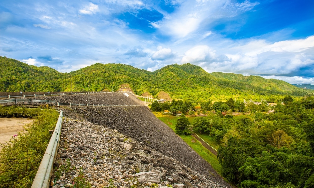 เขื่อนหินที่ยาวเหยียดทอดผ่านหุบเขาเขียวชอุ่ม มีภูเขาที่ปกคลุมด้วยป่าอยู่เบื้องหลัง—ทัศนียภาพอันงดงามสำหรับผู้ที่มองหาสถานที่ท่องเที่ยวในกาญจนบุรีท่ามกลางธรรมชาติและความสงบเงียบ.