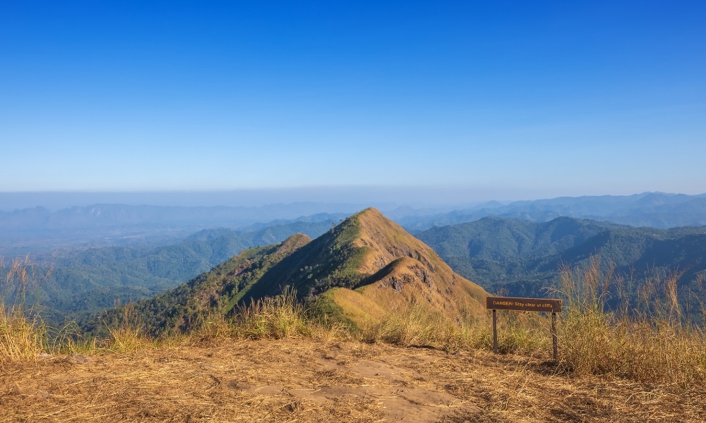 ยอดเขาที่ล้อมรอบด้วยเนินเขาสีเขียวภายใต้ท้องฟ้าสีฟ้าใส มีหญ้าแห้งและป้ายไม้ในเบื้องหน้า—เหมาะสำหรับกา รเที่ยวกาญจนบุรี และสำรวจความงามตามธรรมชาติของสถานที่ท่องเที่ยวในกาญจนบุรี.