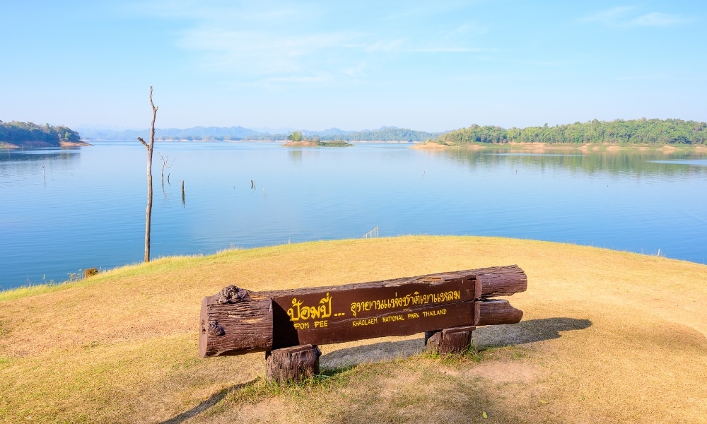 ป้ายไม้ที่มีตัวอักษรภาษาไทยตั้งอยู่บนชายฝั่งหญ้าที่มองเห็นทะเลสาบสงบและภูเขาไกล ๆ ภายใต้ท้องฟ้าสีฟ้าใส เชิญชวนให้คุณมาเที่ยวกาญจนบุรี.