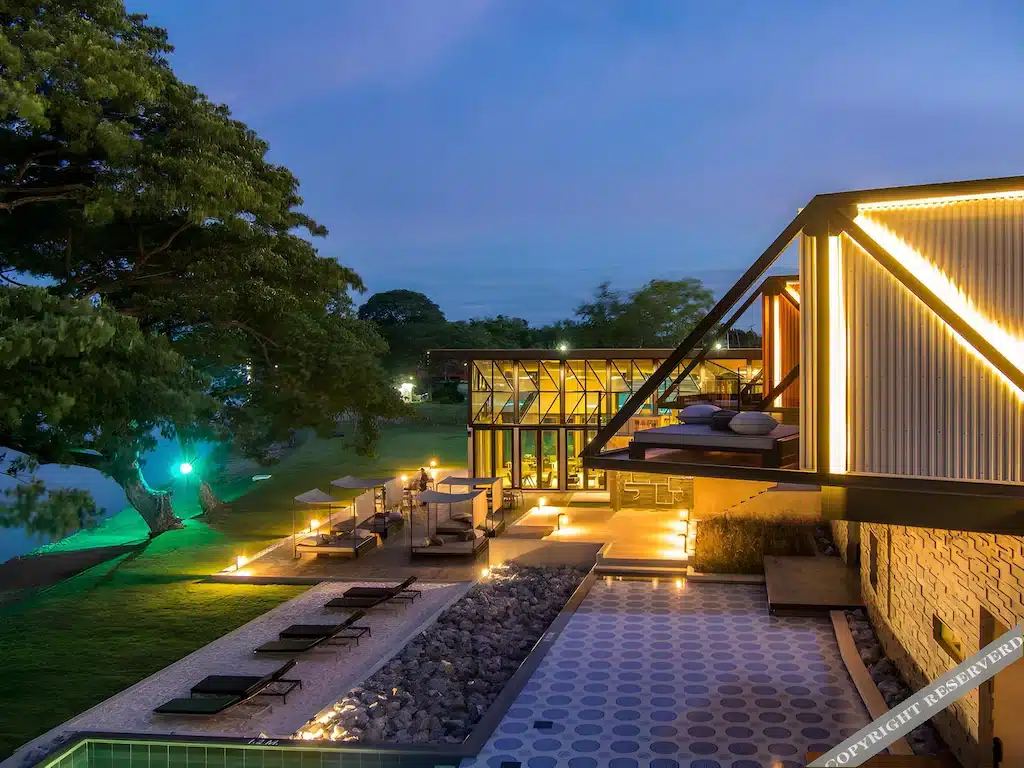 บ้านสมัยใหม่พร้อมหน้าต่างบานใหญ่ สระว่ายน้ำ เก้าอี้อาบแดด และไฟภายนอกในสวนที่จัดแต่งอย่างสวยงามในยามเย็น—เหมาะสำหรับการพักผ่อนหลังจากสำรวจสถานที่ท่องเที่ยวในกาญจนบุรี.
