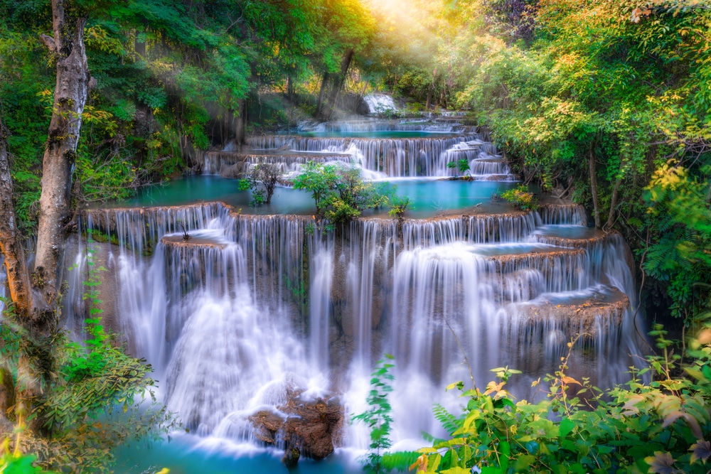 น้ำตกหลายชั้นไหลลดหลั่นผ่านป่าเขียวชอุ่มใน กาญจนบุรีที่เที่ยว โดยมีแสงแดดส่องผ่านต้นไม้ สร้างบรรยากาศธรรมชาติที่เงียบสงบ เหมาะสำหรับผู้ที่ต้องการ เที่ยวกาญจนบุรี.