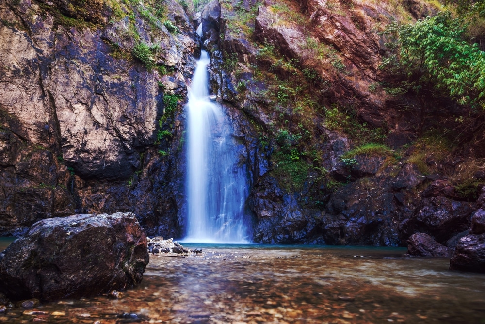 น้ำตกขนาดเล็กไหลลดหลั่นลงมาตามหน้าผาหินสู่แอ่งน้ำตื้นใสสะอาด ล้อมรอบด้วยโขดหินและพืชพรรณสีเขียวบางตา—เป็นสถานที่อันงดงามราวกับภาพวาดในบรรดาแหล่งท่องเที่ยวในกาญจนบุรี เหมาะสำหรับผู้ที่ต้องการท่องเที่ยวและสัมผัสธรรมชาติอย่างแท้จริง.