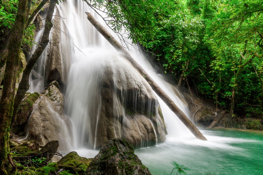 น้ำตกไหลลดหลั่นลงมาตามโขดหินลงสู่แอ่งน้ำสีฟ้าเทอร์ควอยซ์ที่โอบล้อมด้วยต้นไม้เขียวชอุ่ม มีท่อนไม้ล้มพาดอยู่ริมโขดหิน—เหมาะสำหรับผู้ที่มองหาสถานที่ท่องเที่ยวในกาญจนบุรีท่ามกลางความงดงามของธรรมชาติ.