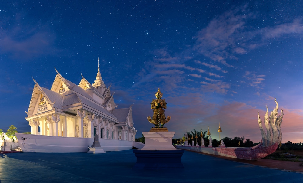 วัดไทยสีขาวที่มีรายละเอียดหลังคาอันวิจิตรตั้งอยู่ใต้ท้องฟ้าที่เต็มไปด้วยดวงดาวในหนึ่งในสถานที่ท่องเที่ยวที่ดีที่สุดของกาญจนบุรี พร้อมด้วยรูปปั้นทองคำและประติมากรรมงูนาคา.