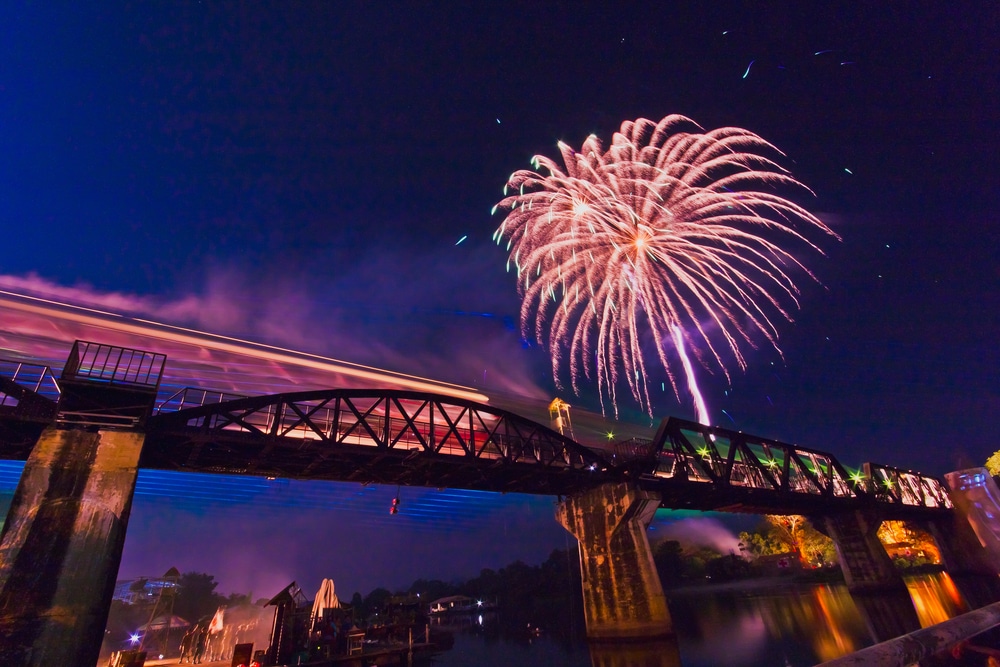 รถไฟข้ามสะพานเหล็กในเวลากลางคืน ณ หนึ่งใน สถานที่ท่องเที่ยวชื่อดังของกาญจนบุรี ขณะที่ดอกไม้ไฟหลากสีสันระเบิดขึ้นเหนือ สะท้อนแสงระยิบระยับในน้ำเบื้องล่าง.