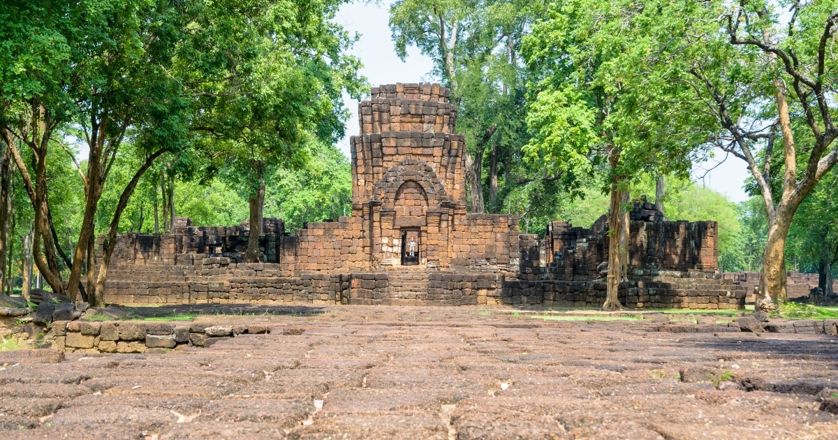 ซากปรักหักพังของวัดหินโบราณที่ล้อมรอบด้วยต้นไม้เขียวขจี มีซุ้มประตูตรงกลางและทางเดินหินที่สึกกร่อนตามกาลเวลา—สถานที่ที่ไม่ควรพลาดใน กาญจนบุรีที่เที่ยว สำหรับผู้ที่ชื่นชอบประวัติศาสตร์.