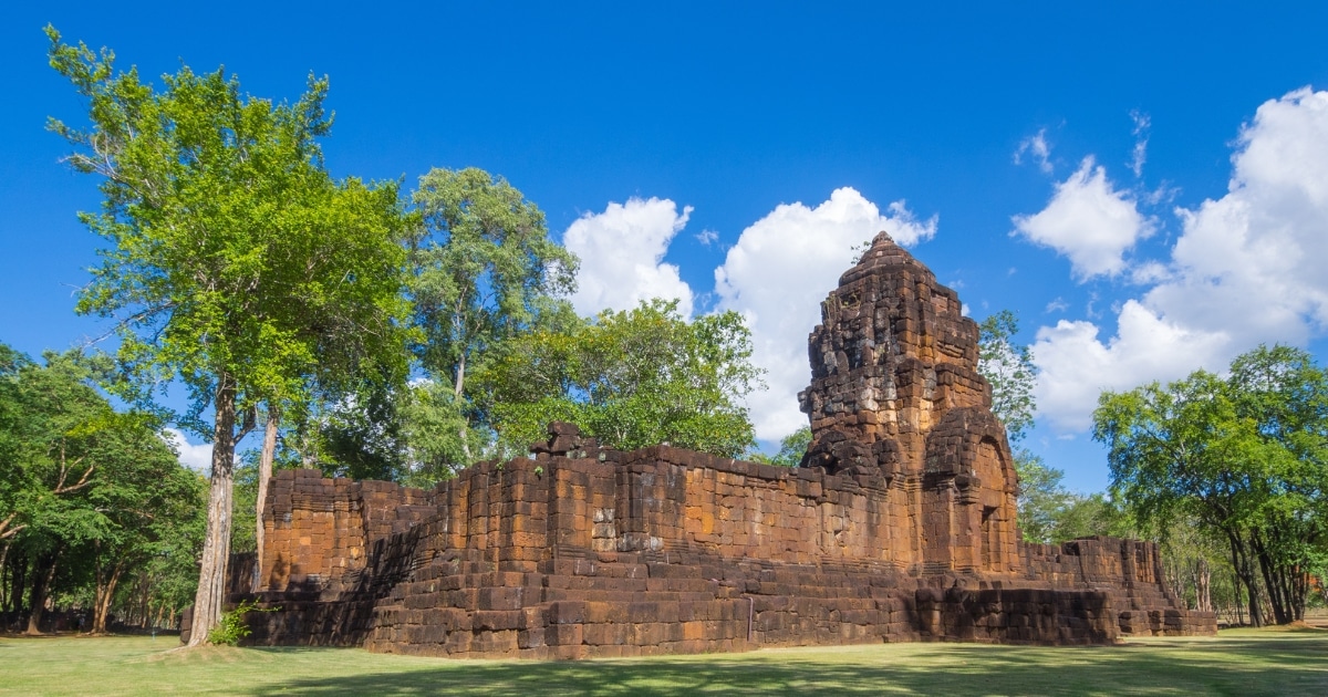 ซากวัดหินโบราณที่ล้อมรอบด้วยต้นไม้เขียวขจีและหญ้าภายใต้ท้องฟ้าสีฟ้าสดใสที่มีเมฆกระจายอยู่ นี่คือ สถานที่ท่องเที่ยวกาญจนบุรี ที่ต้องไปเยือนในกาญจนบุรีสำหรับผู้ที่ต้องการสำรวจประวัติศาสตร์อันรุ่มรวยของภูมิภาคนี้.