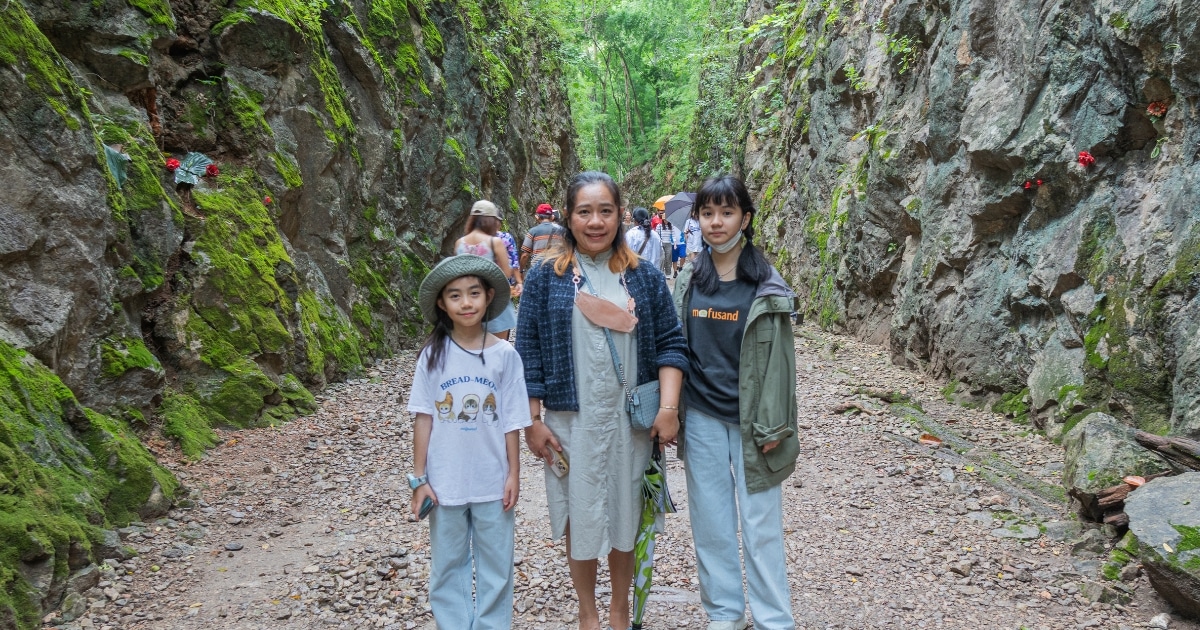 คนสามคนยืนอยู่บนเส้นทางหินระหว่างหน้าผาที่ปกคลุมด้วยมอส โดยมีคนอื่นๆ เดินนำหน้าไปใต้ร่มไม้—ถ่ายทอดความงดงามตามธรรมชาติของ สถานที่ท่องเที่ยวในกาญจนบุรี .