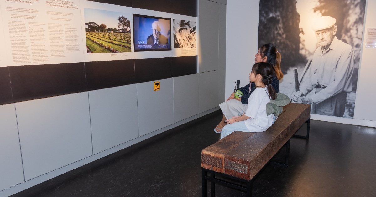 สองคนนั่งอยู่บนม้านั่งในพิพิธภัณฑ์ในกาญจนบุรีที่เที่ยว กำลังดูวิดีโอที่ผนังพร้อมภาพถ่ายทางประวัติศาสตร์และแผ่นป้ายข้อความอยู่ข้างๆ.รวม ที่เที่ยวกาญจนบุรี คัดสถานที่สวยๆ บรรยากาศฟินๆ โอบล้อมด้วยขุนเขาเขียวขจีแห่งภาคตะวันตก มีแม่น้ำสายสำคัญอย่าง “แม่น้ำแคว” ไหลผ่านกลางเมืองสวยๆ