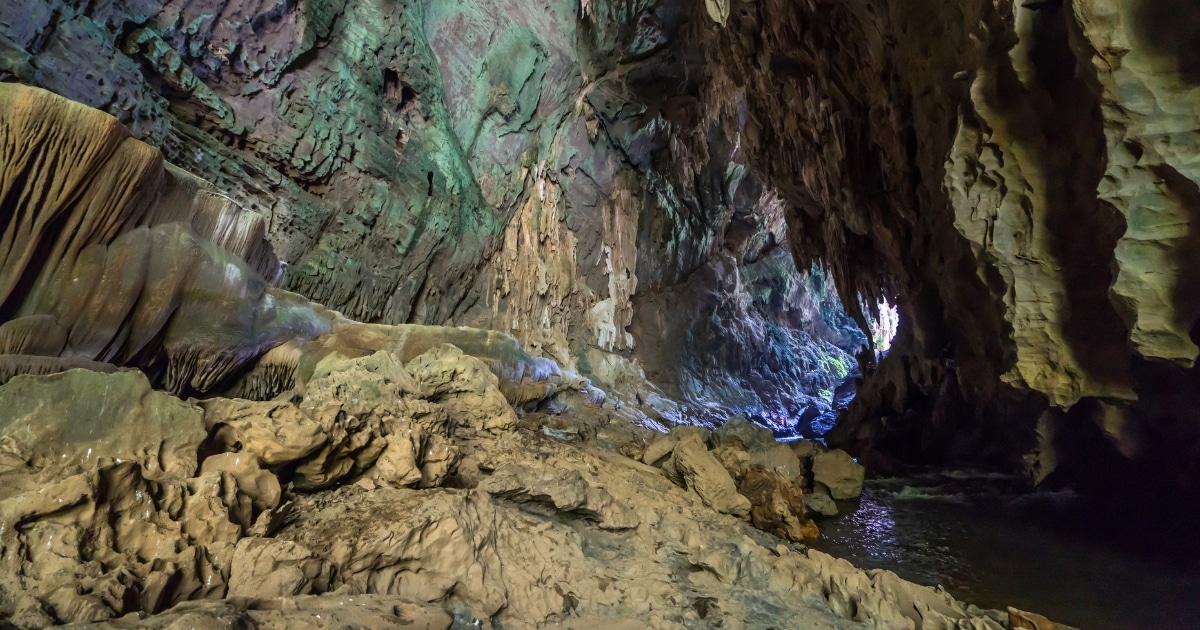 ภายในถ้ำหินที่มีผนังขรุขระ หินย้อย และลำธารเล็กๆ ที่ไหลไปยังช่องเปิดซึ่งมีแสงแดดส่องเข้ามา—อัญมณีที่ซ่อนอยู่ท่ามกลางสถานที่ท่องเที่ยวในกาญจนบุรี.