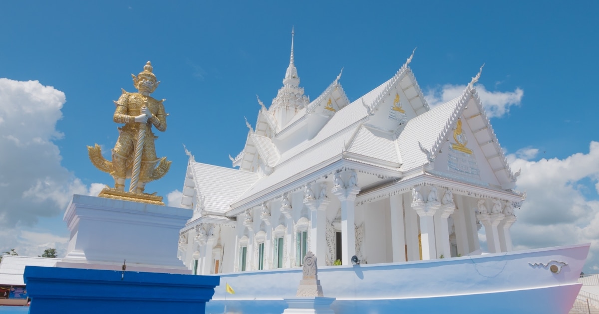 วัดสีขาวที่มียอดแหลมประดับประดาอย่างวิจิตรตั้งอยู่ใต้ท้องฟ้าสีคราม พร้อมด้วยรูปปั้นทองคำในเบื้องหน้า—ตัวอย่างอันงดงามของสถานที่ท่องเที่ยวในกาญจนบุรีที่รอให้คุณไปค้นพบ.