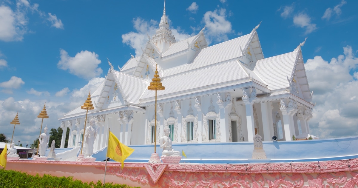 วัดสีขาวประดับประดาอย่างวิจิตรพร้อมลวดลายแกะสลักและรูปปั้นที่ซับซ้อน ล้อมรอบด้วยธงสีเหลืองและรั้วตกแต่งสีชมพูใต้ท้องฟ้าสีคราม—หนึ่งในสถานที่ท่องเที่ยวในกาญจนบุรีที่ไม่ควรพลาดสำหรับนักท่องเที่ยวที่ชื่นชอบการเที่ยวชมสถานที่ในกาญจนบุรี.