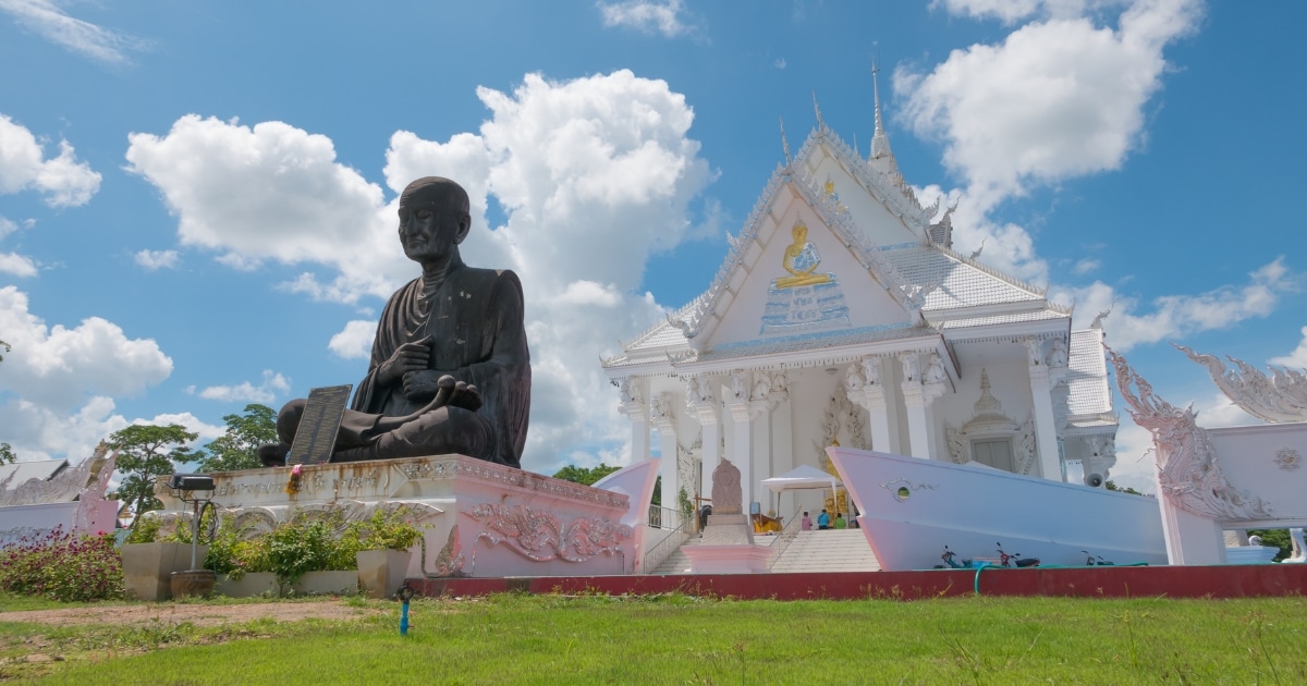 รูปปั้นขนาดใหญ่สีดำนั่งอยู่หน้าวัดสีขาวประดับประดาอย่างสวยงาม ท้องฟ้าสีฟ้าและเมฆลอยอยู่เบื้องบน ทุ่งหญ้าและดอกไม้สีชมพูแซมอยู่เบื้องหน้า ทำให้เป็นสถานที่ท่องเที่ยวที่ไม่ควรพลาดสำหรับนักท่องเที่ยวที่ต้องการความสวยงามและวัฒนธรรมของกาญจนบุรี.